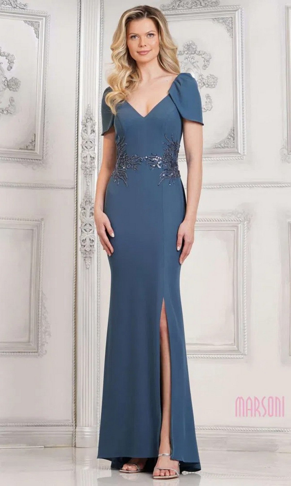 A woman stands indoors wearing the Marsoni MV1297, a floor-length blue sheath formal dress with short sleeves, beaded waist, thigh-high slit, and open-toe heels.